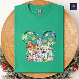 May include: A green t-shirt featuring a colorful graphic of Disney's Toy Story characters. The design includes Woody, Buzz Lightyear, and other characters, set against a castle and palm trees. The shirt is displayed on a wooden plate.