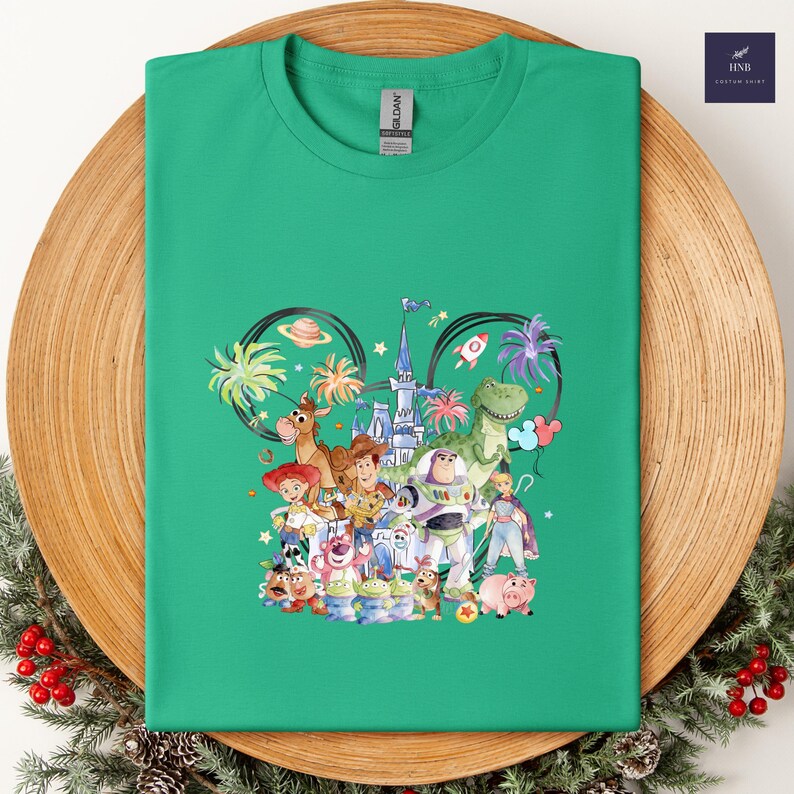 May include: A green t-shirt featuring a colorful graphic of Disney's Toy Story characters. The design includes Woody, Buzz Lightyear, and other characters, set against a castle and palm trees. The shirt is displayed on a wooden plate.