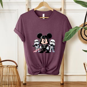 Pu&ograve; includere: T-shirt bordeaux con un disegno a cartoni animati di Topolino vestito da Darth Vader, affiancato da due Stormtrooper. Mickey tiene una spada laser rossa. Il disegno &egrave; centrato sulla parte anteriore della maglietta.