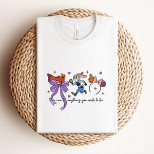 May include: White t-shirt featuring a cartoon illustration of a fox and a rabbit running, with a purple bow and a pair of mouse ears. The text "You can be anything you wish to be" is below the illustration.