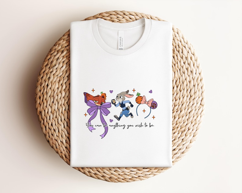 May include: White t-shirt featuring a cartoon illustration of a fox and a rabbit running, with a purple bow and a pair of mouse ears. The text "You can be anything you wish to be" is below the illustration.