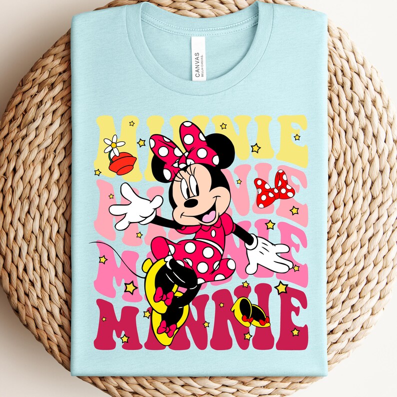 May include: Light blue t-shirt featuring a graphic of Minnie Mouse in a red polka dot dress and bow. The word "MINNIE" is displayed in a retro font with yellow, pink, and red colors. Small stars and a flower graphic are also included.
