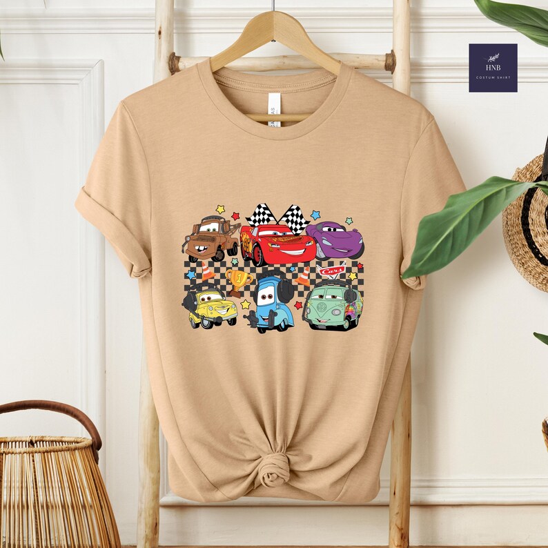 May include: Tan t-shirt featuring a colorful graphic of cartoon cars. The design includes a red race car with a checkered flag, a purple car, a blue car, a yellow car, a brown car, and a green van. The shirt is hanging on a wooden hanger.