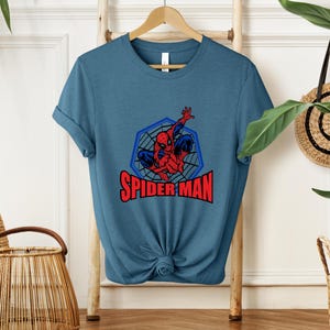 May include: A blue t-shirt featuring a graphic of Spider-Man in a web-like design. The superhero is depicted in red and blue, with the words "SPIDER-MAN" in red block letters below the image. The shirt is hanging on a wooden hanger.