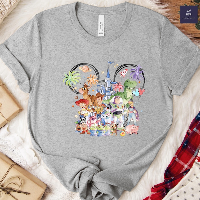 May include: A gray t-shirt featuring a colorful graphic of Disney's Toy Story characters inside a Mickey Mouse ear outline. The shirt has the text "CANVAS" on the upper left side.