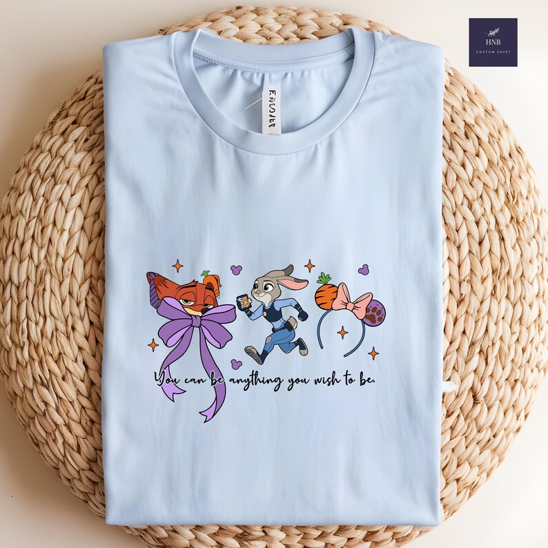 May include: Light blue t-shirt featuring a cartoon design with a fox, a rabbit, and a carrot-shaped headband. The shirt includes the text "You can be anything you wish to be."