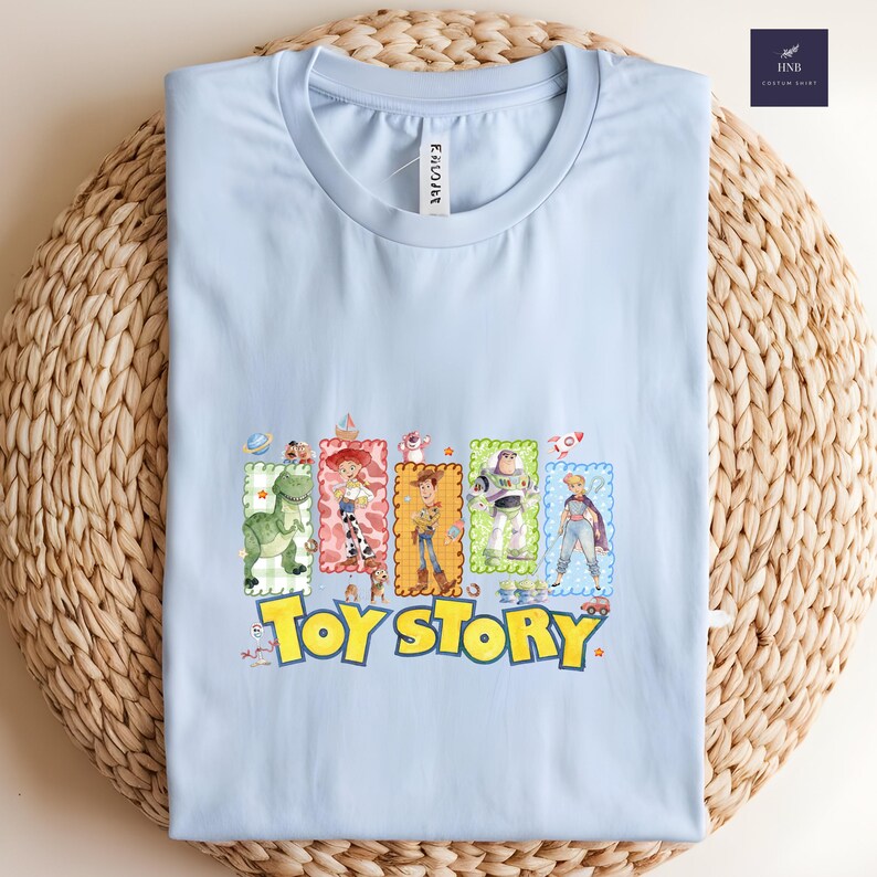 Puede incluir: Una camiseta azul claro con un gr&aacute;fico colorido de personajes de la pel&iacute;cula animada "Toy Story". El dise&ntilde;o incluye im&aacute;genes de un dinosaurio, una vaquera, un vaquero, un guardabosques espacial y m&aacute;s, con las palabras "Toy Story" en amarillo.