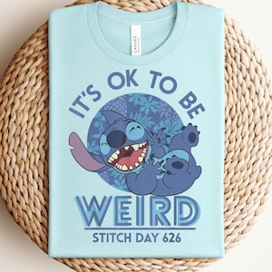 Puede incluir: Camiseta azul claro con un dise&ntilde;o del personaje de dibujos animados Stitch y el texto "IT'S OK TO BE WEIRD" y "STITCH DAY 626". El gr&aacute;fico incluye un c&iacute;rculo azul con motivos florales. La camiseta es de un material suave.