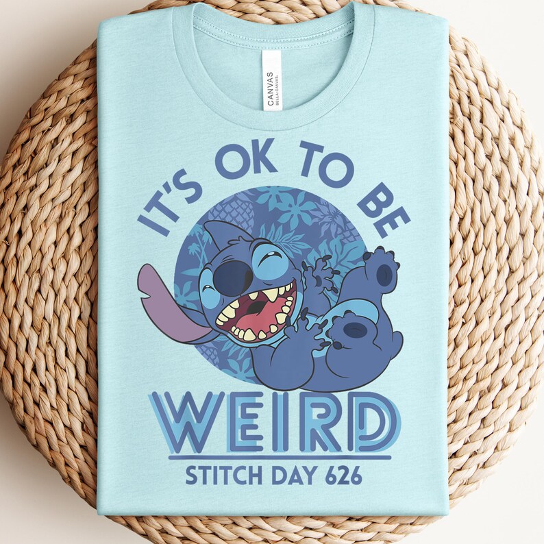 Puede incluir: Camiseta azul claro con un dise&ntilde;o del personaje de dibujos animados Stitch y el texto "IT'S OK TO BE WEIRD" y "STITCH DAY 626". El gr&aacute;fico incluye un c&iacute;rculo azul con motivos florales. La camiseta es de un material suave.