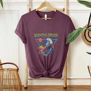 May include: A maroon t-shirt with a graphic of characters from the Mandalorian series. The design includes the text "Mandalorian" in a rainbow gradient, a triangular frame, and images of the characters.