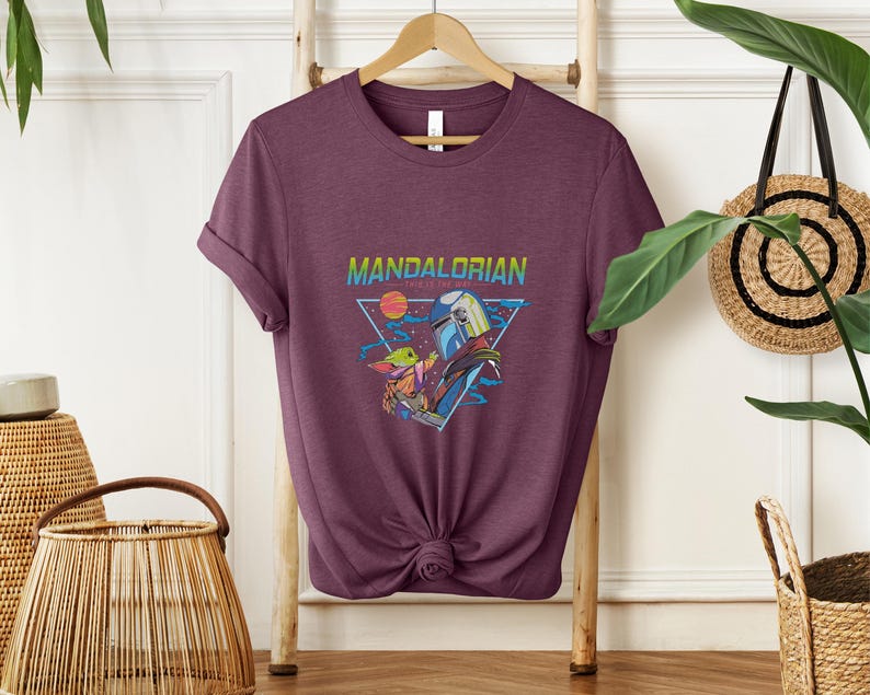 May include: A maroon t-shirt with a graphic of characters from the Mandalorian series. The design includes the text "Mandalorian" in a rainbow gradient, a triangular frame, and images of the characters.
