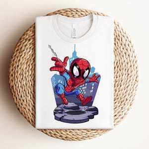May include: White t-shirt featuring a cartoon illustration of Spider-Man in a dynamic pose. The superhero is depicted in his classic red and blue suit, with a cityscape backdrop. The shirt is folded on a woven, circular surface.