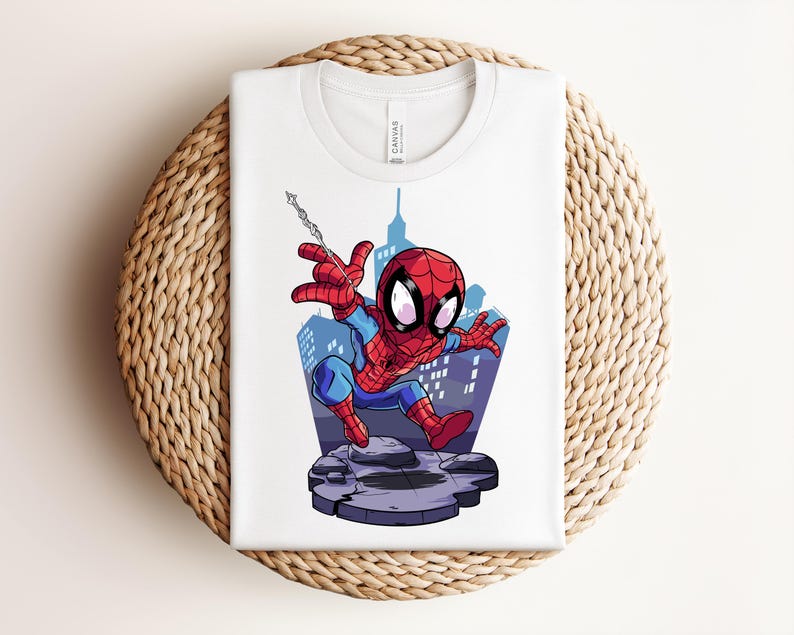May include: White t-shirt featuring a cartoon illustration of Spider-Man in a dynamic pose. The superhero is depicted in his classic red and blue suit, with a cityscape backdrop. The shirt is folded on a woven, circular surface.