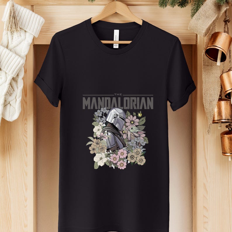 Puede incluir: Camiseta negra con un gr&aacute;fico de The Mandalorian y un dise&ntilde;o floral. El texto "THE MANDALORIAN" est&aacute; impreso encima de la imagen. La camiseta est&aacute; colgada en una percha de madera.