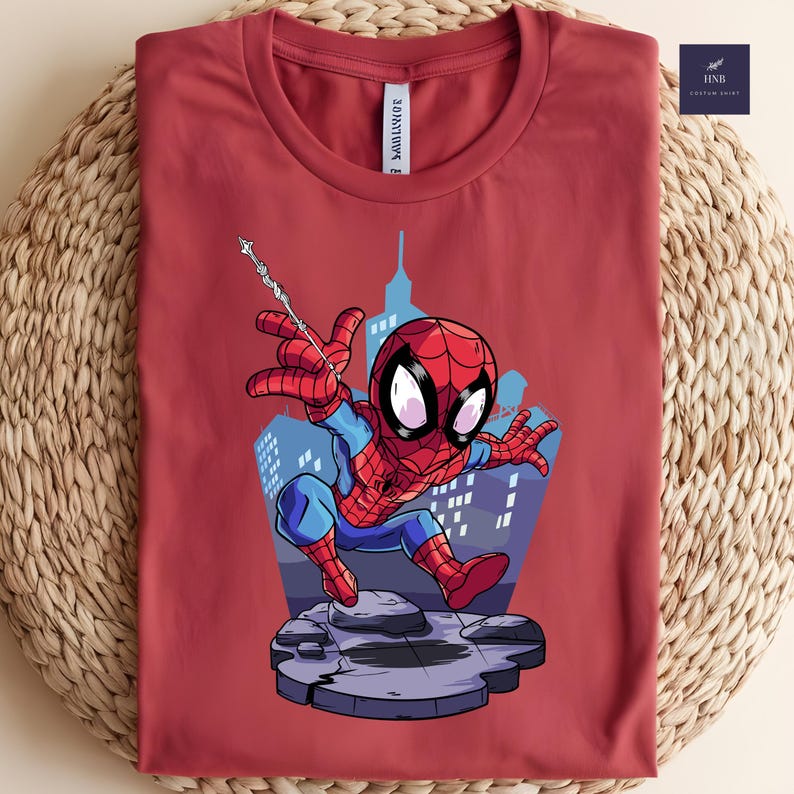 May include: A maroon t-shirt featuring a cartoon Spider-Man graphic. The superhero is depicted in a dynamic pose with a web, set against a cityscape backdrop. The shirt has a crew neck and the text "30ADMIRE" is printed on the neck.