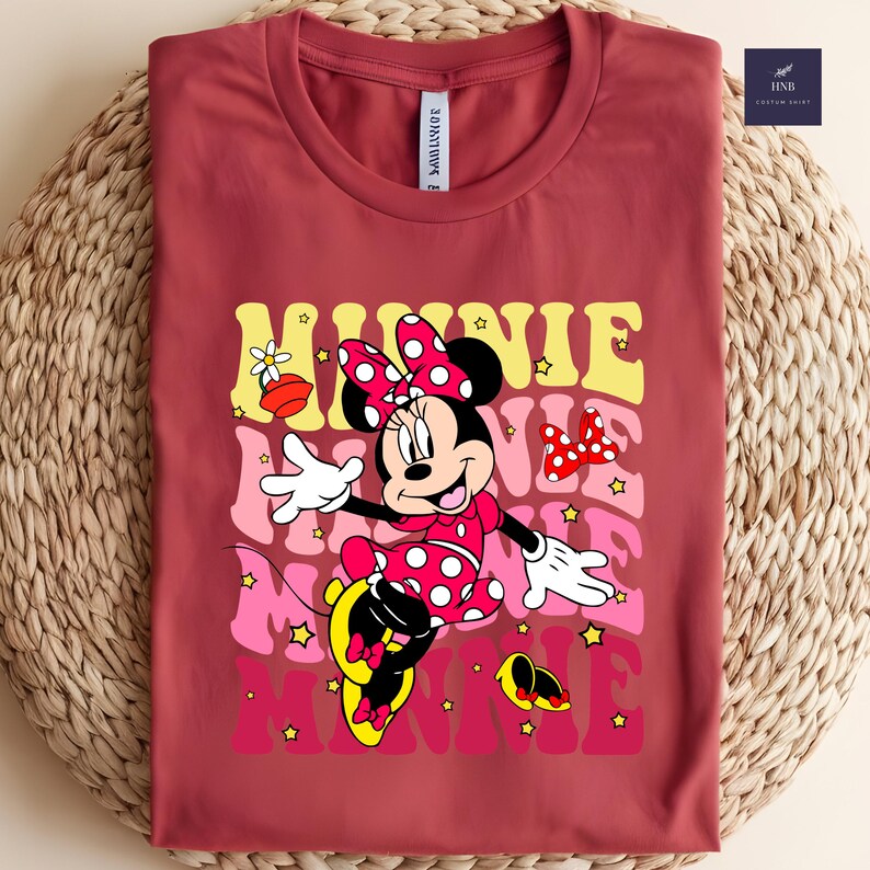 May include: A maroon t-shirt featuring a graphic of Minnie Mouse in a red polka-dot dress and bow. The word "Minnie" is layered in yellow, pink, and red, with star accents. The shirt is folded on a woven surface.