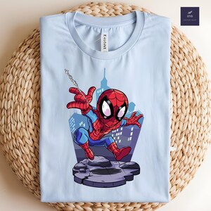 May include: Light blue t-shirt featuring a cartoon Spider-Man graphic. The superhero is depicted in his classic red and blue suit, with a web slung from his hand. The background shows a cityscape. The shirt is folded on a woven surface.