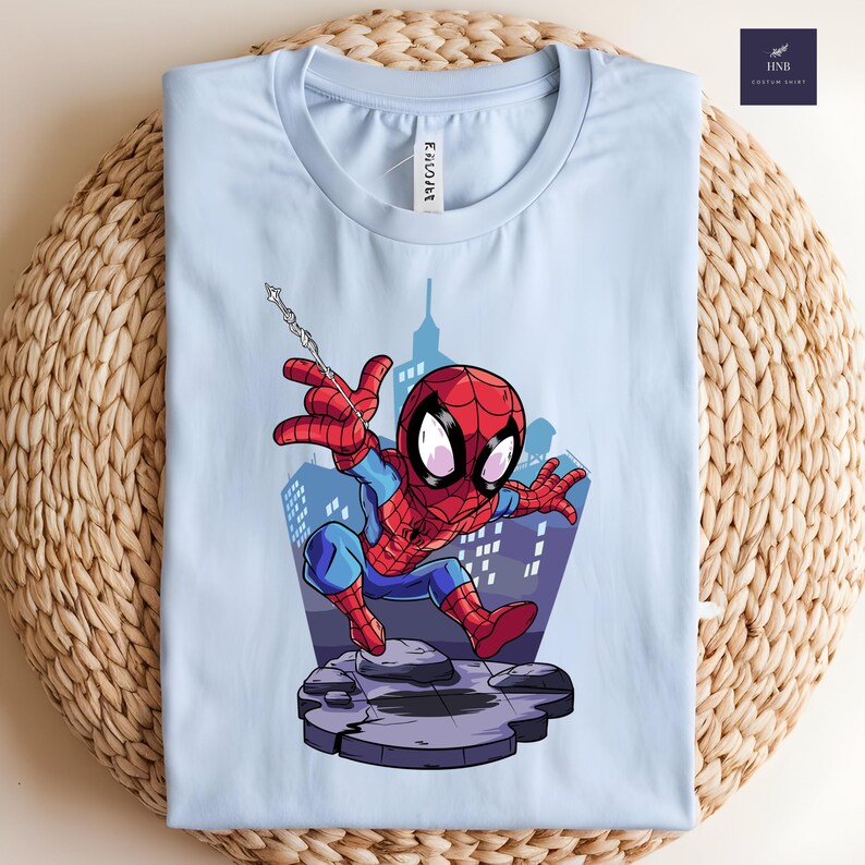 May include: Light blue t-shirt featuring a cartoon Spider-Man graphic. The superhero is depicted in his classic red and blue suit, with a web slung from his hand. The background shows a cityscape. The shirt is folded on a woven surface.