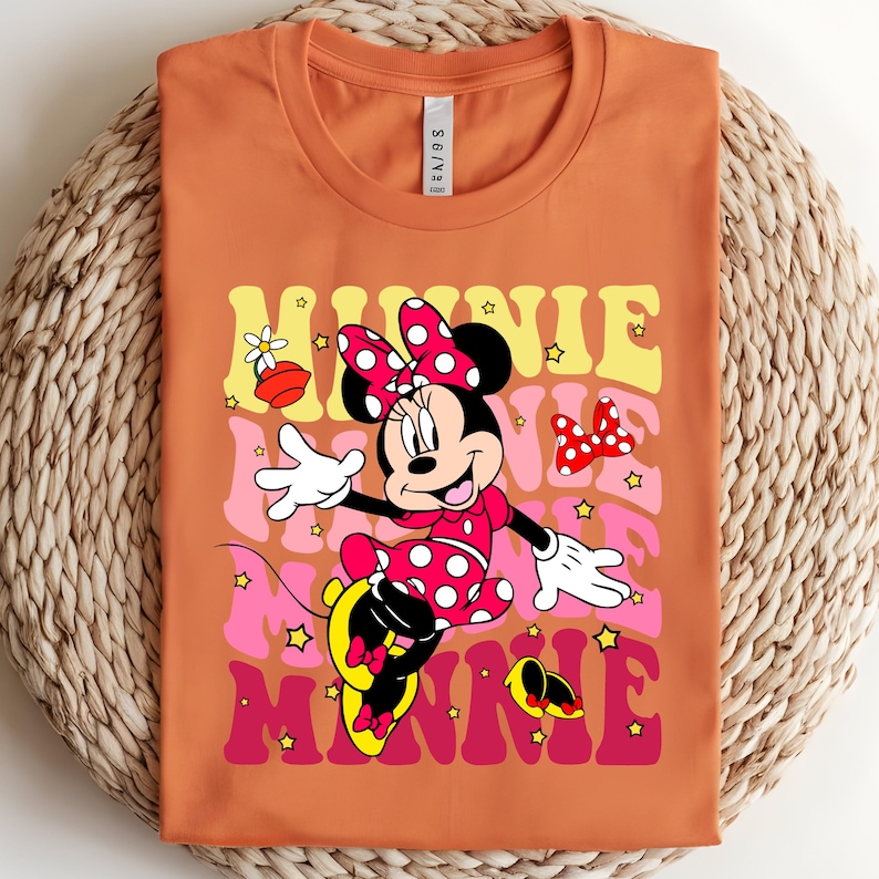 May include: Orange t-shirt featuring Minnie Mouse in a retro design. The design includes the word "MINNIE" in layered colors of yellow, pink, and red, with stars and Minnie Mouse in a polka dot dress and bow.