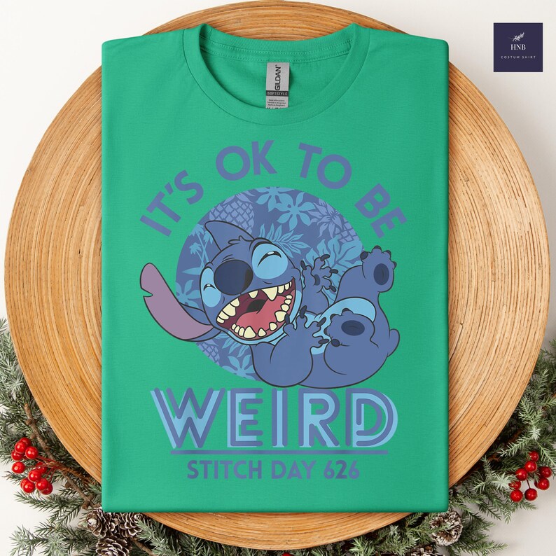 Puede incluir: Camiseta verde con un dise&ntilde;o de dibujos animados de Stitch y el texto "IT'S OK TO BE WEIRD" y "STITCH DAY 626". La imagen muestra un personaje Stitch azul con un fondo circular p&uacute;rpura.