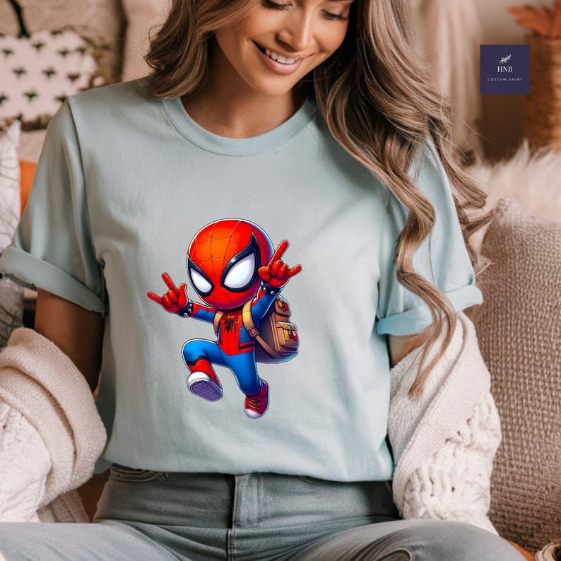 Puede incluir: Camiseta azul claro con un dise&ntilde;o de Spider-Man de dibujos animados. El personaje lleva un traje rojo y azul, una m&aacute;scara roja y ojos blancos, y una peque&ntilde;a mochila. La camiseta tiene las mangas dobladas.