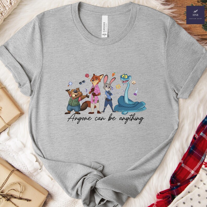 K&ouml;nnte beinhalten: Hellgraues T-Shirt mit einer Cartoon-Illustration von Zootopia-Charakteren. Das Design zeigt einen Biber, einen Fuchs, ein Kaninchen und eine Schlange mit dem Text "Anyone can be anything". Das T-Shirt ist aus weichem Material.