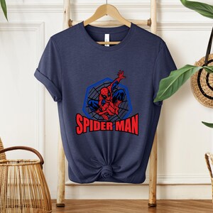 May include: Navy blue t-shirt featuring a Spider-Man graphic. The design includes a red and blue Spider-Man figure within a blue web-like hexagon, with the words "SPIDER MAN" in red. The shirt is knotted at the bottom.
