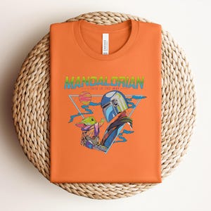 May include: Orange t-shirt with a graphic of The Mandalorian and Grogu, with the text "MANDALORIAN" in green and blue. The design includes a helmet, a baby Yoda-like character, and a geometric background. The shirt is folded on a woven mat.
