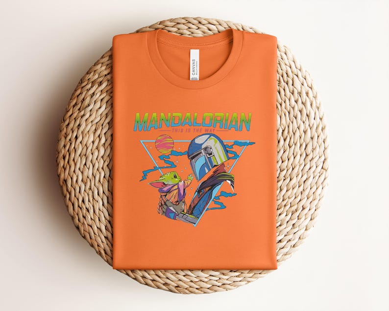 May include: Orange t-shirt with a graphic of The Mandalorian and Grogu, with the text "MANDALORIAN" in green and blue. The design includes a helmet, a baby Yoda-like character, and a geometric background. The shirt is folded on a woven mat.