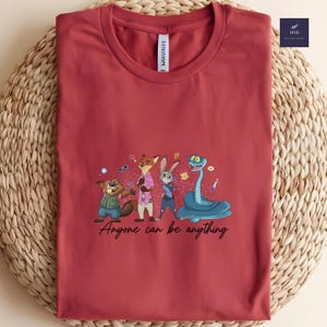 K&ouml;nnte beinhalten: Burgunderfarbenes T-Shirt mit einer Cartoon-Illustration von Tierfiguren, darunter ein Biber, ein Fuchs, ein Kaninchen und eine Schlange. Der Text "Anyone can be anything" steht darunter.