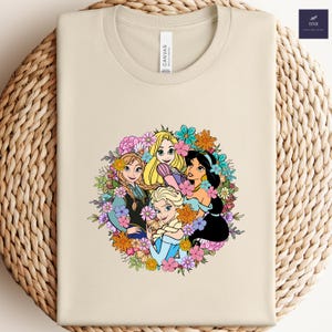 May include: A cream-colored t-shirt featuring a colorful floral design with four animated princess characters. The design includes Anna, Elsa, Rapunzel, and Jasmine, surrounded by a wreath of flowers. The shirt has a crew neck and short sleeves.