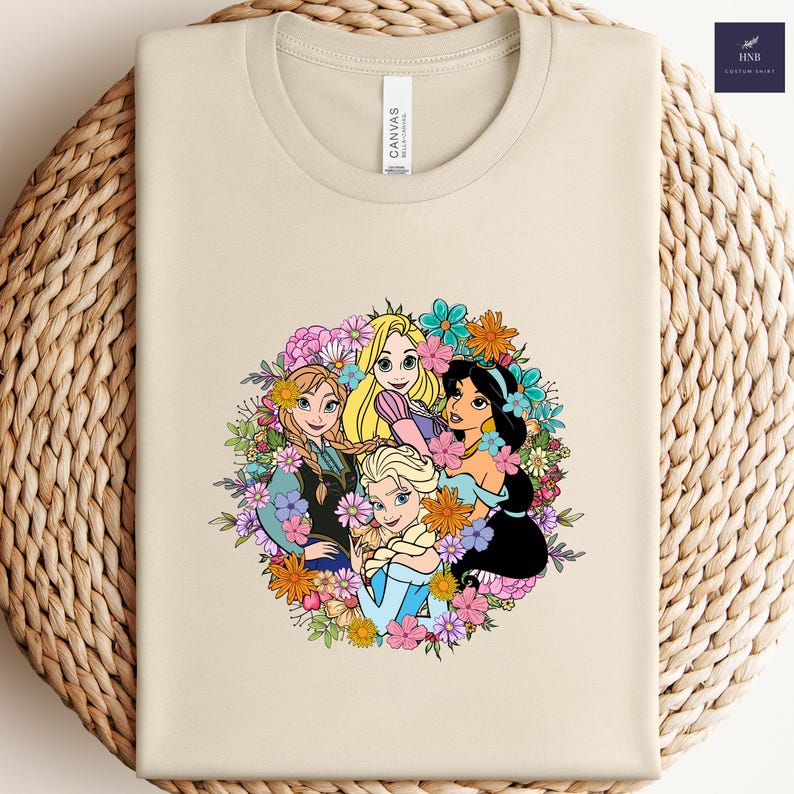 May include: A cream-colored t-shirt featuring a colorful floral design with four animated princess characters. The design includes Anna, Elsa, Rapunzel, and Jasmine, surrounded by a wreath of flowers. The shirt has a crew neck and short sleeves.