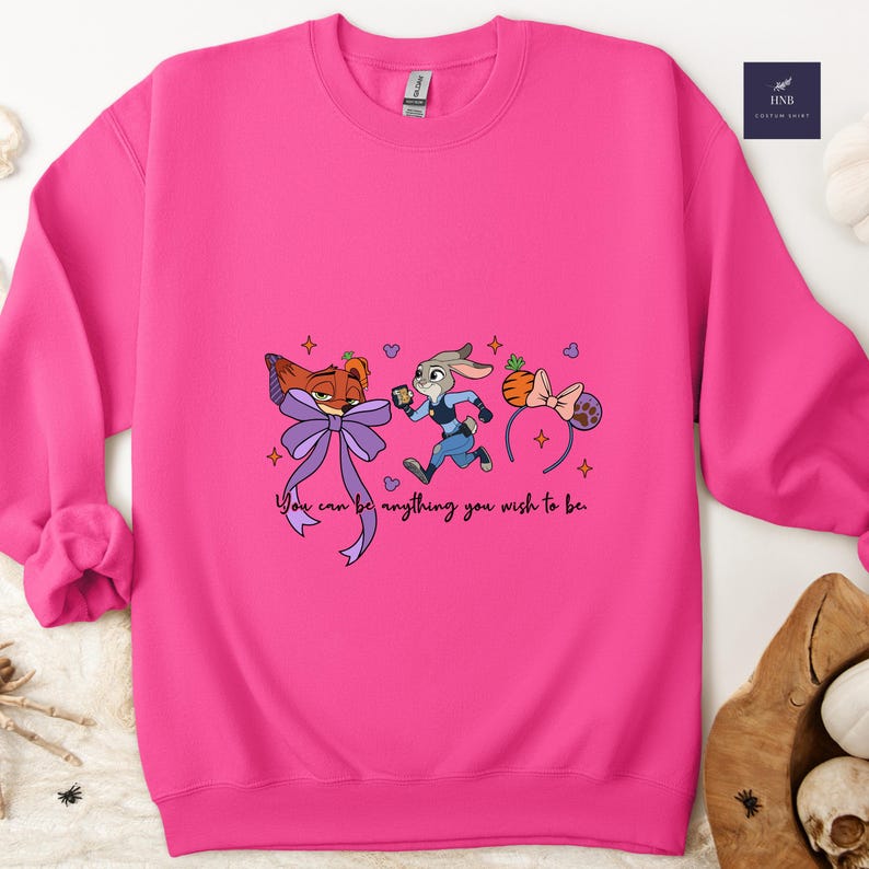 May include: A bright pink sweatshirt featuring cartoon characters from Zootopia. The design includes Nick Wilde with a purple bow, Judy Hopps in a police uniform, and a headband with a carrot and bow. The text "You can be anything you wish to be" is also included.