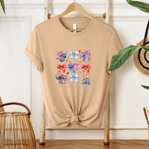 May include: A tan t-shirt featuring a colorful graphic of cartoon characters and bows. The graphic includes blue, pink, and red elements. The shirt is displayed on a wooden hanger.