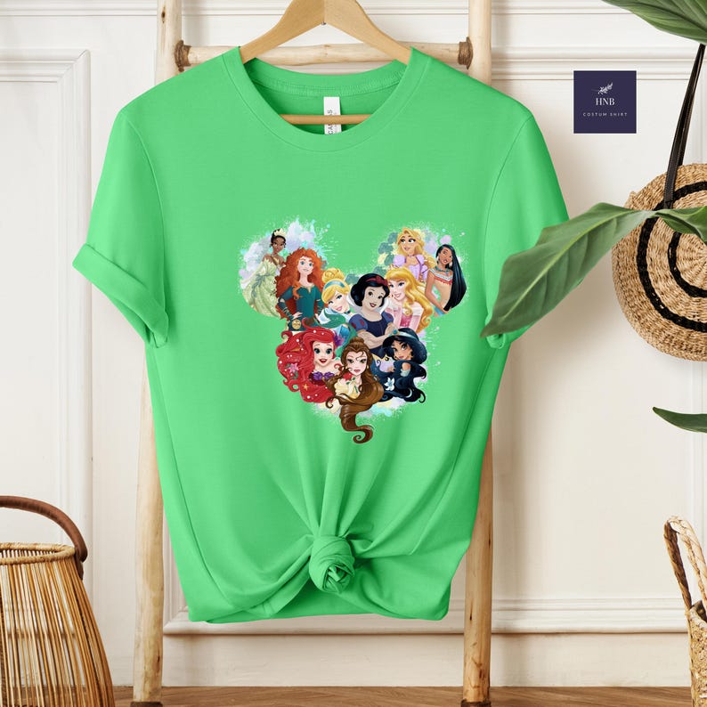 K&ouml;nnte beinhalten: Ein hellgr&uuml;nes T-Shirt mit einem herzf&ouml;rmigen Design, das aus Cartoon-Prinzessinnen besteht. Die Prinzessinnen sind mit ihren charakteristischen Frisuren und Outfits dargestellt. Das T-Shirt hat kurze &Auml;rmel und einen Rundhalsausschnitt.
