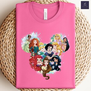 K&ouml;nnte beinhalten: Ein pinkfarbenes T-Shirt mit einem Aufdruck von Disney-Prinzessinnen in Herzform. Das Design zeigt Ariel, Belle, Jasmin, Schneewittchen und andere Prinzessinnen. Das Shirt hat einen Rundhalsausschnitt und ein "Canvas"-Etikett.
