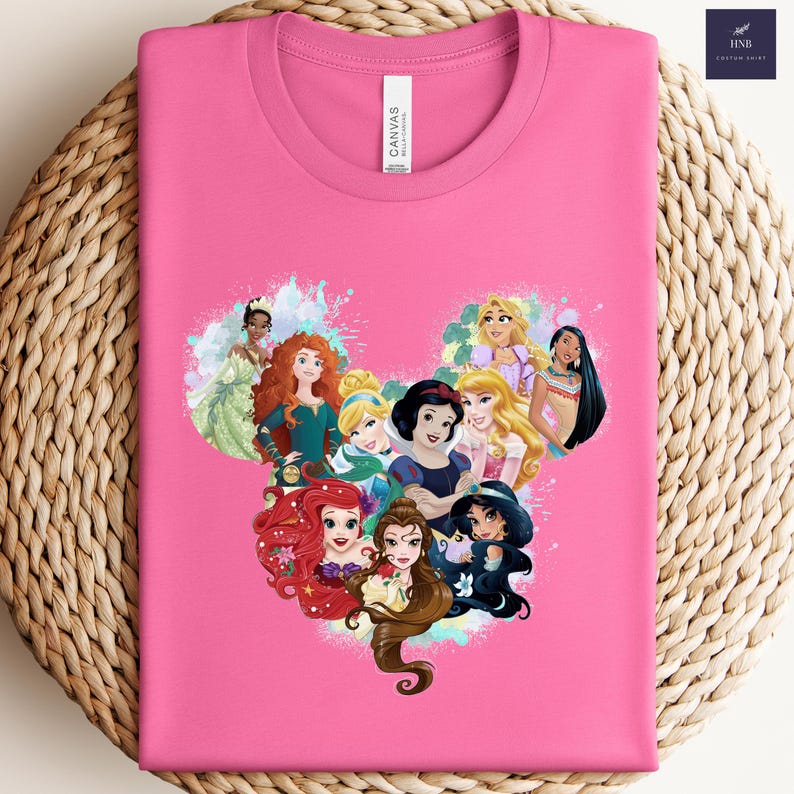 K&ouml;nnte beinhalten: Ein pinkfarbenes T-Shirt mit einem Aufdruck von Disney-Prinzessinnen in Herzform. Das Design zeigt Ariel, Belle, Jasmin, Schneewittchen und andere Prinzessinnen. Das Shirt hat einen Rundhalsausschnitt und ein "Canvas"-Etikett.