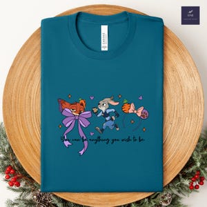 May include: Teal t-shirt featuring cartoon characters and the text "You can be anything you wish to be." The design includes a fox with a purple bow, a running rabbit, and a carrot-shaped headband with a pink bow.