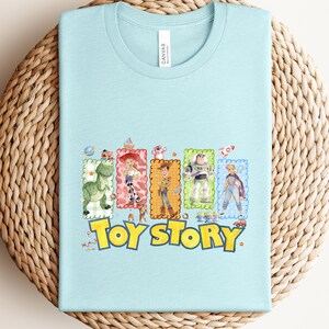 Puede incluir: Camiseta azul claro con un gr&aacute;fico colorido de personajes de la pel&iacute;cula "Toy Story". El gr&aacute;fico incluye im&aacute;genes de un dinosaurio, Woody, Buzz Lightyear y otros personajes, con las palabras "Toy Story" en amarillo.