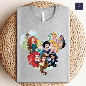 K&ouml;nnte beinhalten: Ein hellgraues T-Shirt mit Rundhalsausschnitt und einem herzf&ouml;rmigen Design aus Cartoon-Prinzessinnen. Das T-Shirt hat ein "CANVAS"-Etikett. Das Design zeigt verschiedene Prinzessinnen.
