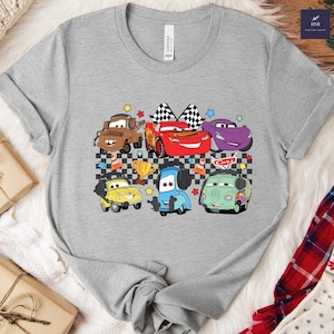 May include: A gray t-shirt featuring a colorful graphic of cartoon cars, including a red race car with checkered flags, a brown truck, and a purple car. The design includes stars, a trophy, and the word "Cars".
