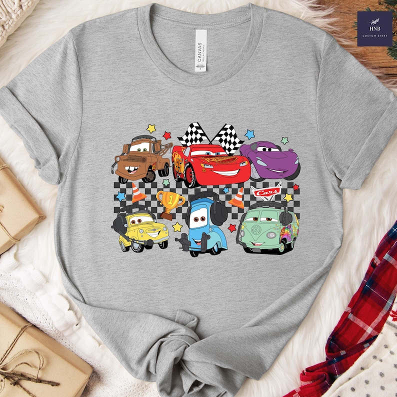 May include: A gray t-shirt featuring a colorful graphic of cartoon cars, including a red race car with checkered flags, a brown truck, and a purple car. The design includes stars, a trophy, and the word "Cars".