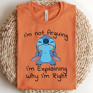 May include: Orange t-shirt featuring a cartoon character, Stitch, with the text "I'm not Arguing, I'm Explaining why I'm Right." The character is blue with large ears and a grumpy expression. The shirt is folded on a woven surface.