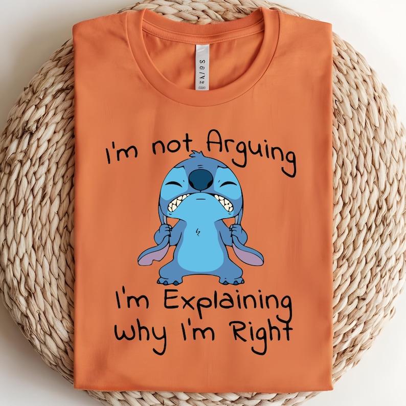 May include: Orange t-shirt featuring a cartoon character, Stitch, with the text "I'm not Arguing, I'm Explaining why I'm Right." The character is blue with large ears and a grumpy expression. The shirt is folded on a woven surface.