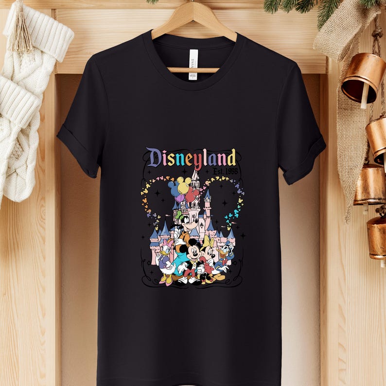 Może przedstawiać: Czarny t-shirt z kr&oacute;tkim rękawem z kolorową grafiką zamku Disneyland i klasycznymi postaciami z kresk&oacute;wek, w tym Myszką Miki, Myszką Minnie, Kaczorem Donaldem, Daisy Duck i Goofym, z nadrukowanym powyżej napisem Disneyland Est. 1955.
