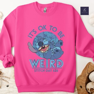 Puede incluir: Una sudadera rosa brillante con un personaje de dibujos animados azul riendo. El texto "IT'S OK TO BE WEIRD" est&aacute; encima del personaje, con "STITCH DAY 626" debajo. El dise&ntilde;o est&aacute; centrado en la parte delantera de la sudadera.