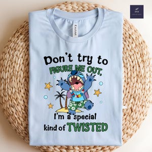 May include: Light blue t-shirt featuring a cartoon character with the text "Don't try to FIGURE ME OUT, I'm a special kind of TWISTED." The graphic includes palm trees, stars, and bubbles. The shirt is folded on a woven surface.