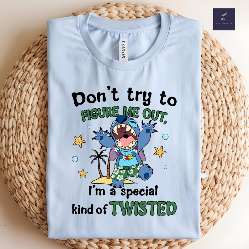 May include: Light blue t-shirt featuring a cartoon character with the text "Don't try to FIGURE ME OUT, I'm a special kind of TWISTED." The graphic includes palm trees, stars, and bubbles. The shirt is folded on a woven surface.