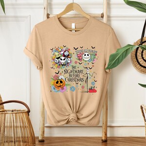 May include: Tan t-shirt featuring a colorful graphic from "The Nightmare Before Christmas." The design includes Jack Skellington, Sally, Zero, and other characters, along with the text "Trick or Treat" and "The Nightmare Before Christmas."