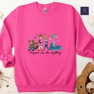 K&ouml;nnte beinhalten: Ein leuchtend pinkfarbenes Sweatshirt mit einer Cartoon-Illustration von Tierfiguren. Das Design zeigt einen Biber, einen Fuchs, ein Kaninchen und eine Schlange mit dem Text "Anyone can be anything" in einer Schreibschrift.